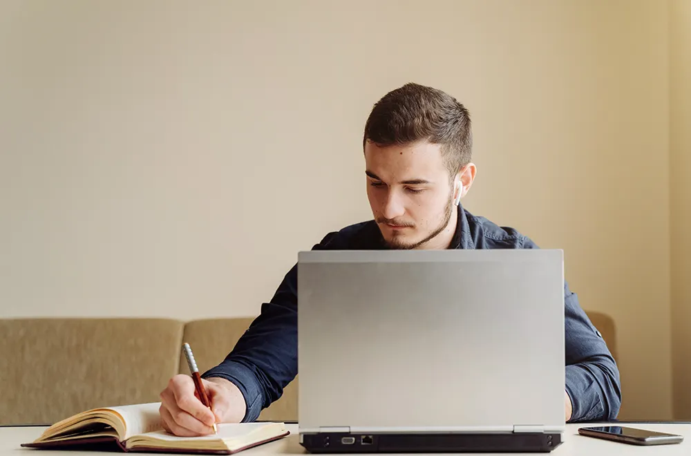 person using his laptop on a desk while making notes on a notepad